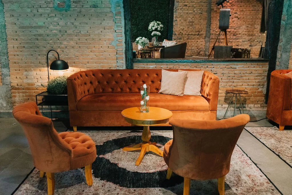 Sala de estar com sofá e cadeiras em veludo de tom laranja, mesa de centro de madeira com uma jarra de flores, parede de tijolos expostos, luminária de mesa acesa, decorativa com plantas e livros.