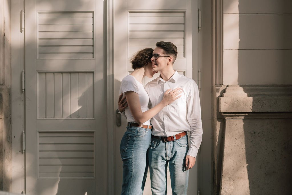 Casal sorridente se abraçando na rua, de frente para uma porta de madeira.