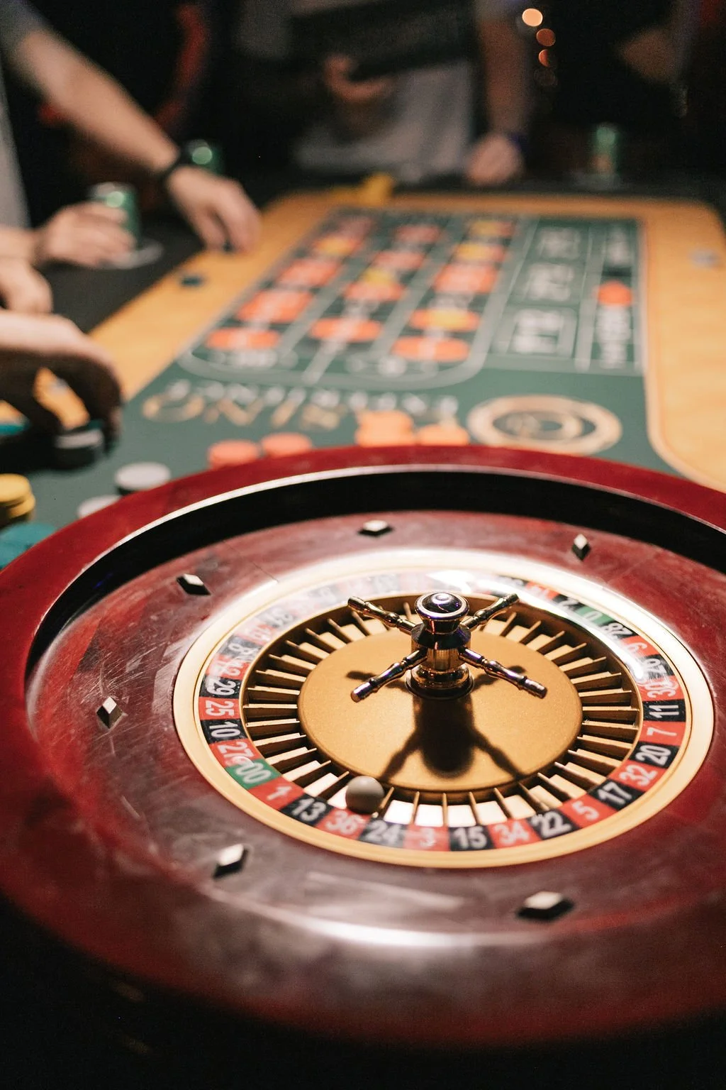 Foco no roleta de um cassino, com mesa de jogo ao fundo e mãos de jogadores ao redor.