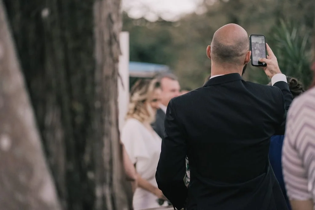 Homem de terno preto tirando uma foto com celular de uma mulher usando máscara facial branca durante um evento ao ar livre.