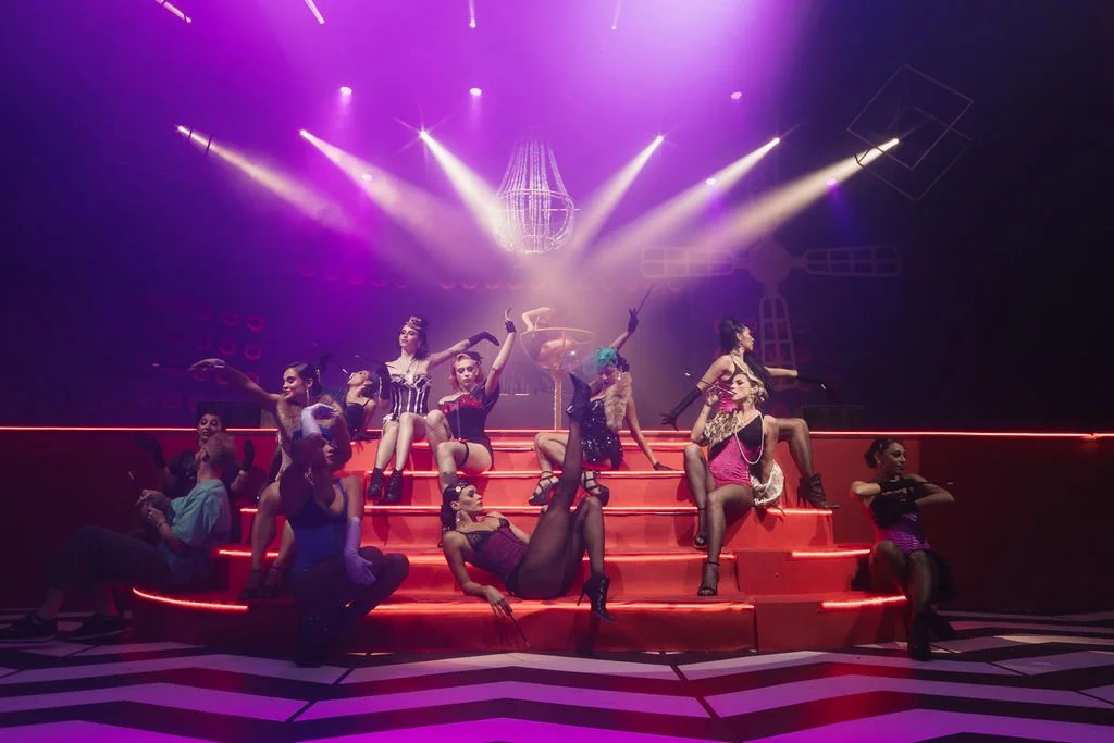 Grupo de pessoas em um palco com luzes coloridas, alguns estão sentados, outros de pé, em roupas de estilo teatral ou de performance, criando uma cena de show ou apresentação artística.