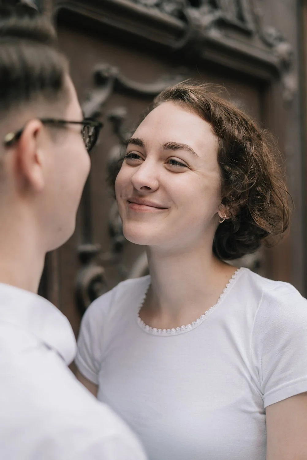 Homem e mulher sorrindo um para o outro em um ambiente interno, com detalhes de uma porta ao fundo.