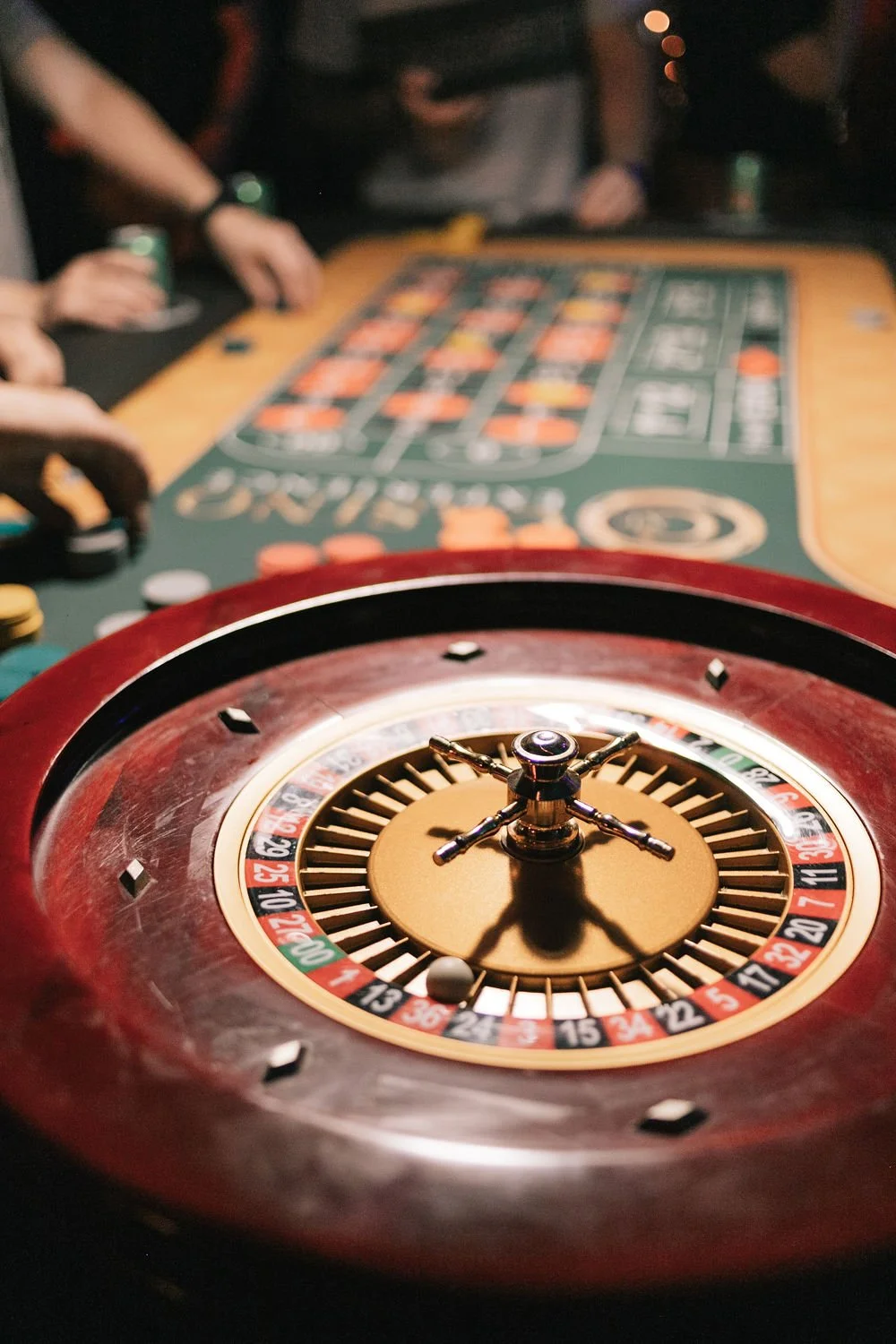 Roleta de cassino com o cursor na posição zero, em primeiro plano, com mesa de roleta e jogadores ao fundo. Ambiente de cassino.