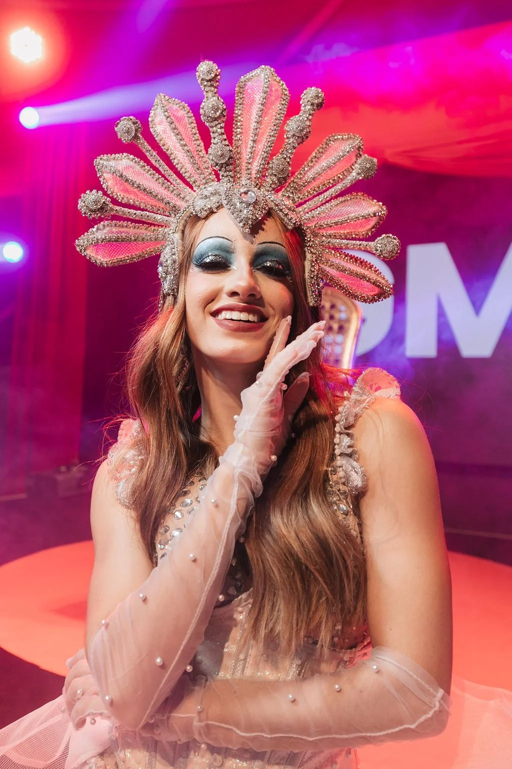 Mulher vestida com fantasia de rainha, usando uma coroa decorada com pérolas e pedras brilhantes, sorrindo em cenário de palco iluminado com luzes coloridas.
