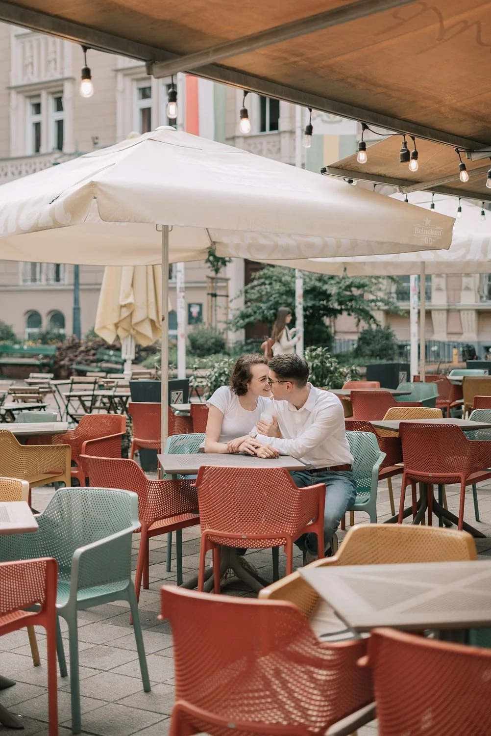 Casal sorridente sentados em uma mesa de um restaurante ao ar livre com cadeiras coloridas sob guarda-sóis e luzes penduradas