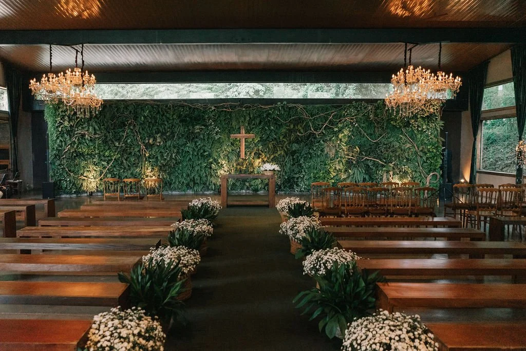 Interior de uma igreja decorada para cerimônia, com bancos de madeira, lustres elegantes, um altar com uma cruz e flores brancas ao longo do corredor central.