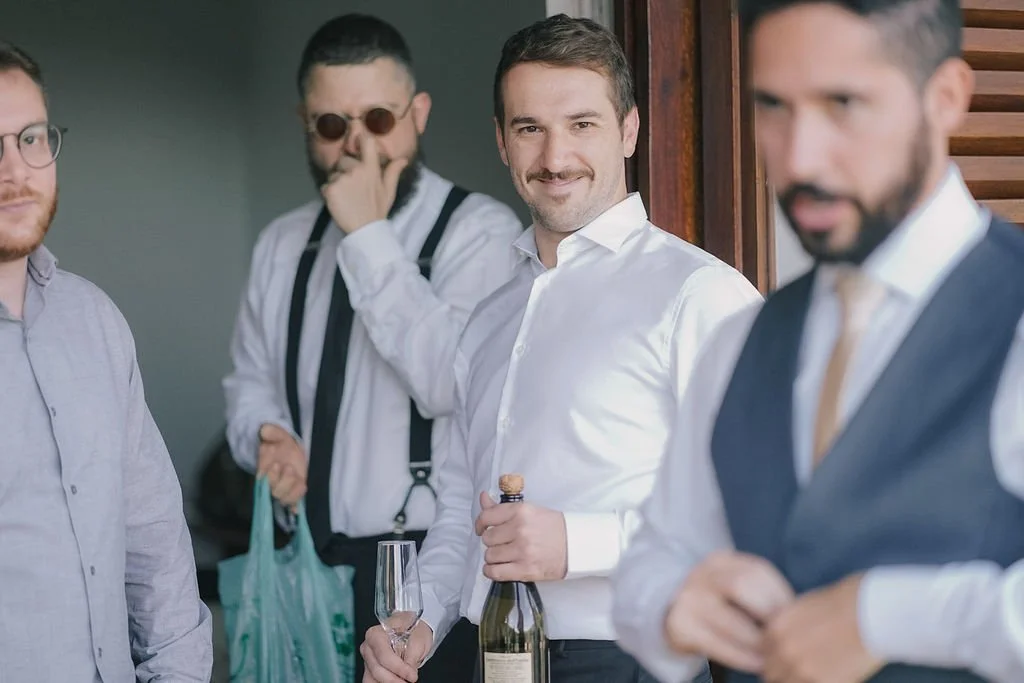 Homem sorridente segurando uma garrafa de espumante e uma taça, rodeado por outros homens em ambiente social