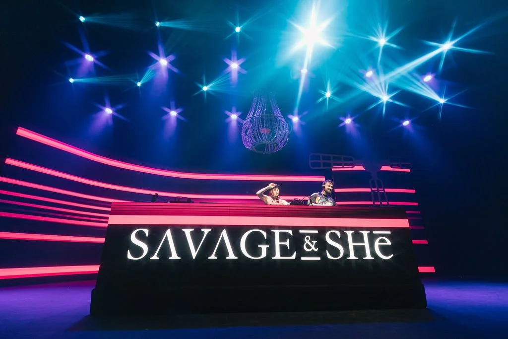 DJ duo Savage & She perform on stage in a nightclub with neon pink and blue lighting, a large LED screen displaying their name, and a chandelier hanging from the ceiling.