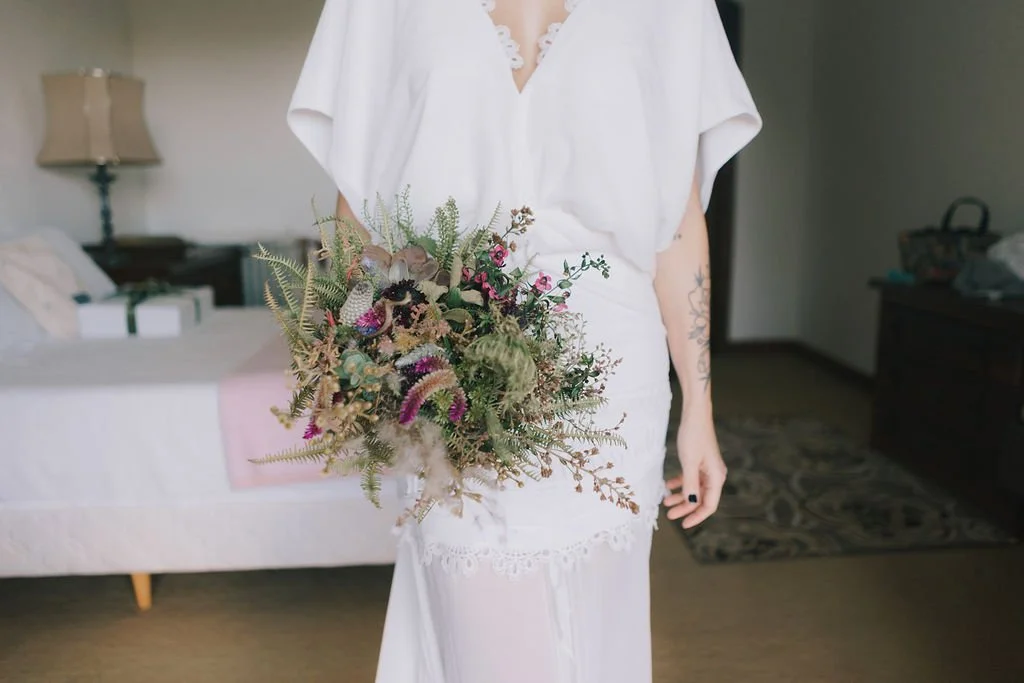 Pessoa vestida de branco segurando um buquê de flores em um quarto.