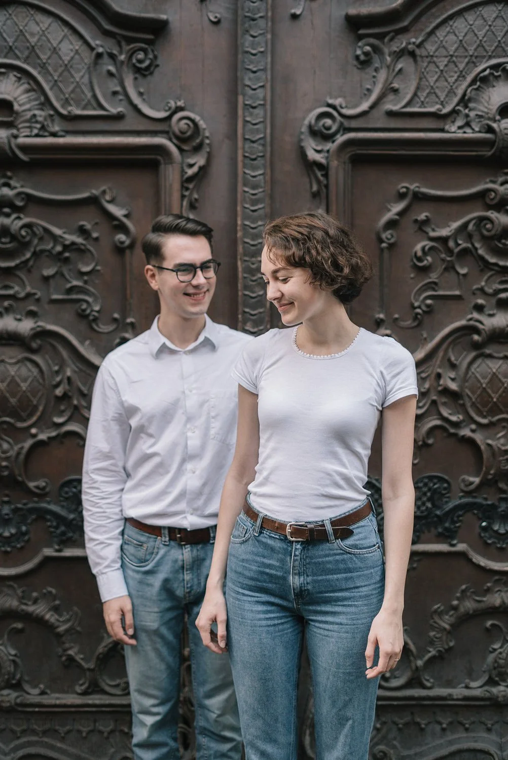 Um homem com óculos e uma mulher com cabelo cacheado, ambos com roupas casuais, em frente a uma porta de madeira detalhada e ornamentada.