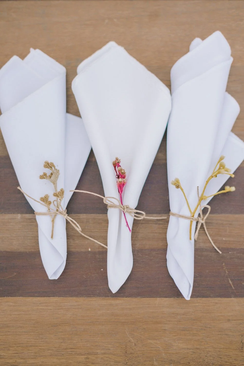 Três pequenos ramos de flores secos, cada um envolto em papel branco e amarrado com barbante, dispostos sobre uma mesa de madeira.