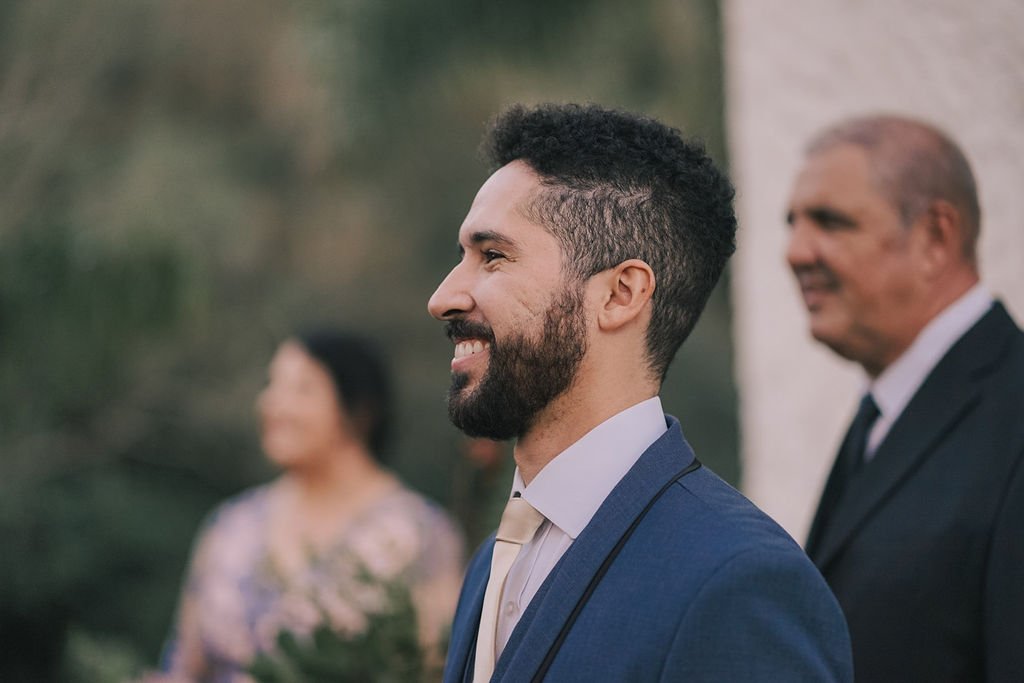 Homem sorridente vestindo terno azul, ao lado de um homem mais velho. Fundo desfocado com uma mulher visível também ao fundo.