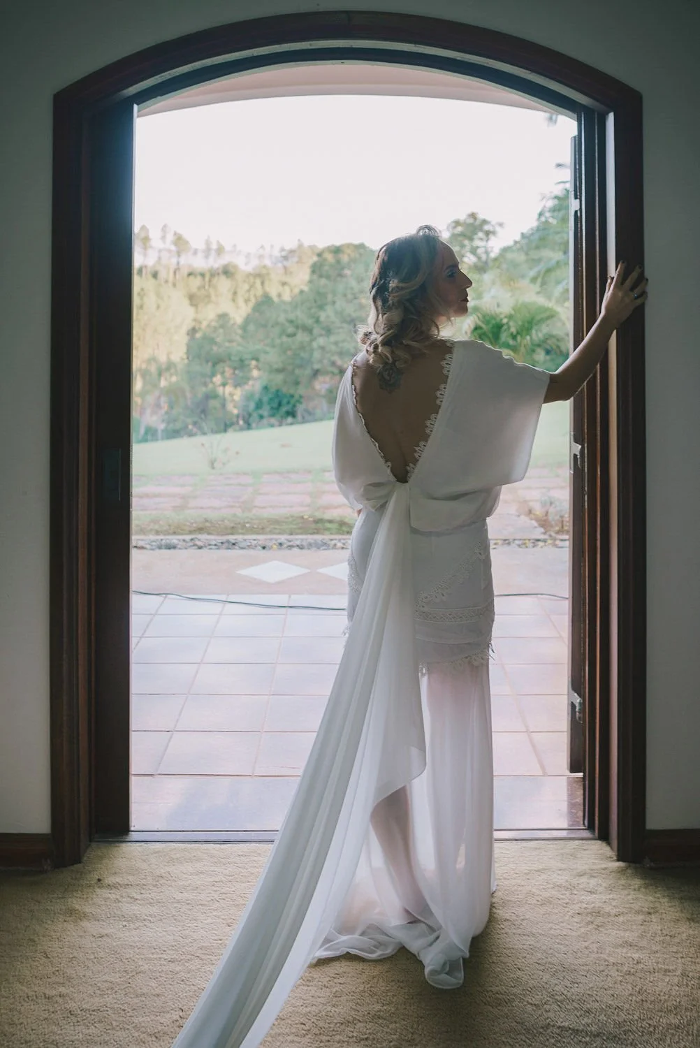 Mulher em um vestido branco com decote nas costas, de cabelo cacheado, de pé na porta de uma casa, olhando para fora, com vista para uma área verde com árvores ao fundo.
