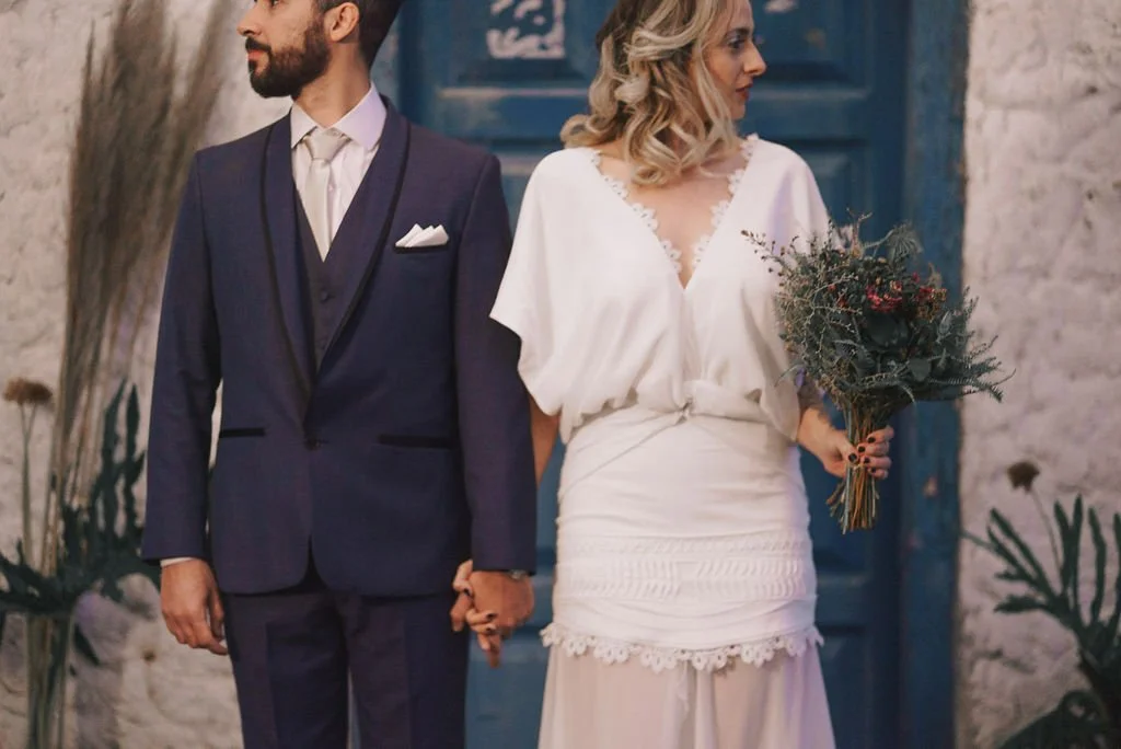 Casal de noivos segurando as mãos, com o noivo vestindo terno azul escuro e a noiva vestindo vestido branco com flores no cabelo e segurando um buquê de flores.