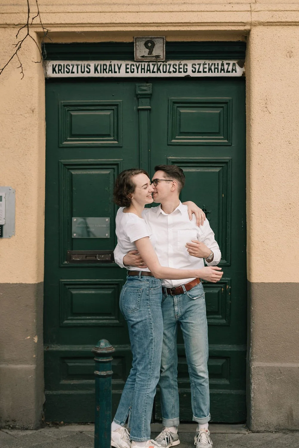 Casal sorridente se abraçando na frente de uma porta verde, com uma placa em húngaro acima dela