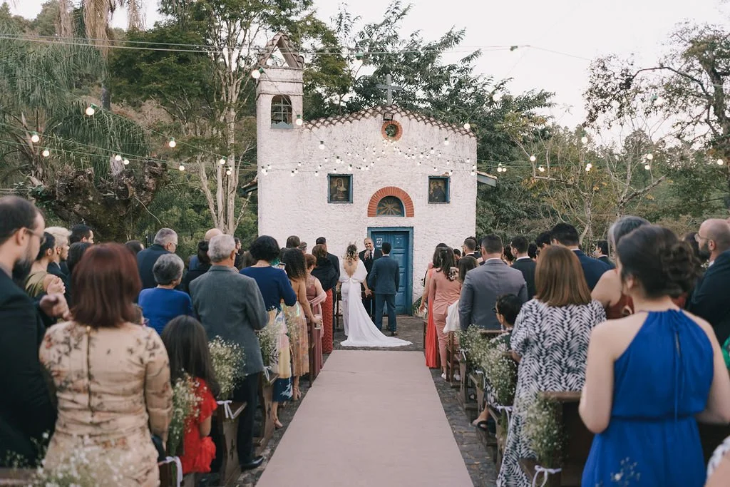 Casamento ao ar livre em frente a uma pequena igreja branca, com convidados assentos de ambos os lados do caminho de entrada, árvores ao redor, luzes decorativas e o casal de noivos na frente, de mãos dadas.