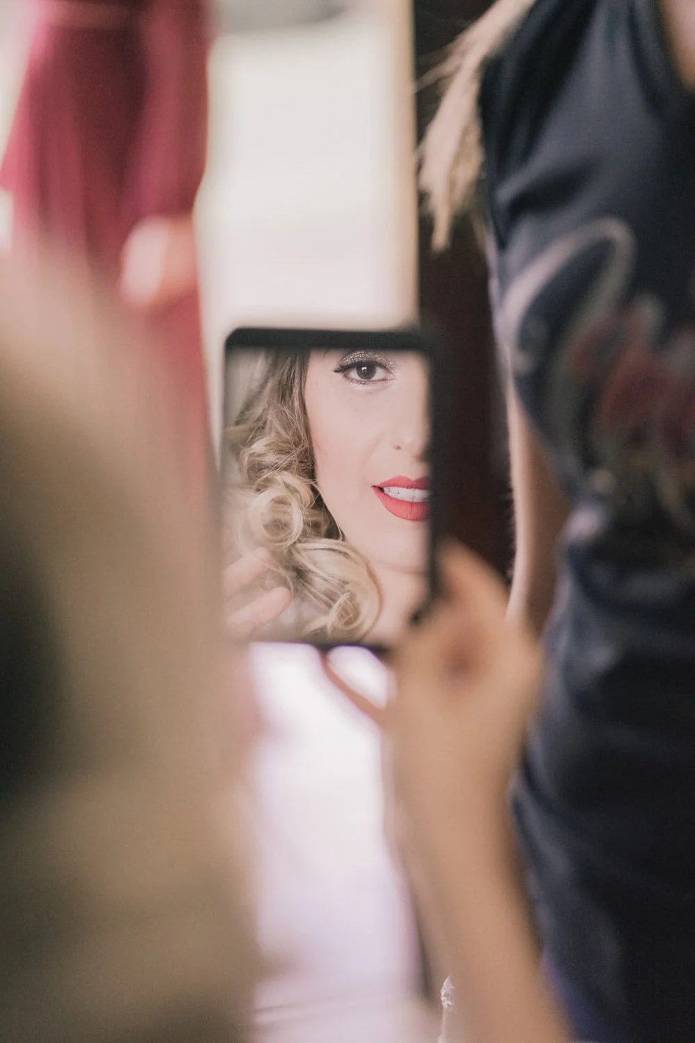 Mulher com cabelo cacheado loiro, maquiagem marcante, sorrindo levemente e usando batom vermelho, sendo vista parcialmente através de um espelho de mão em um ambiente interno