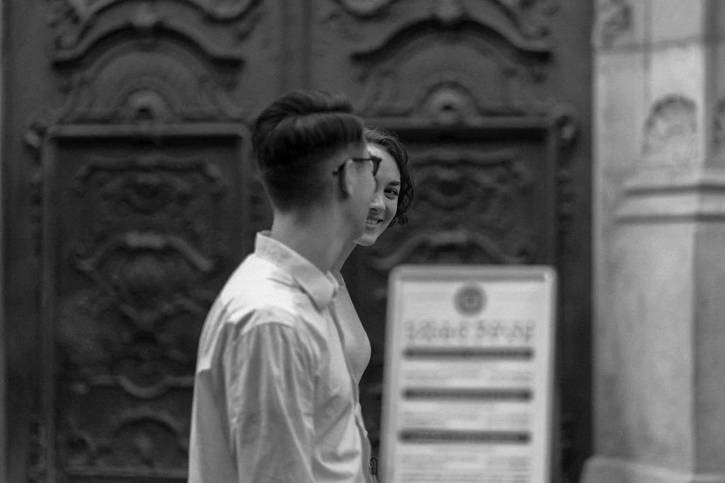 Duas pessoas conversando em frente a uma porta decorada, uma mulher sorri para uma pessoa de óculos e cabelo curto, ambas de costas, em uma parede de pedra.