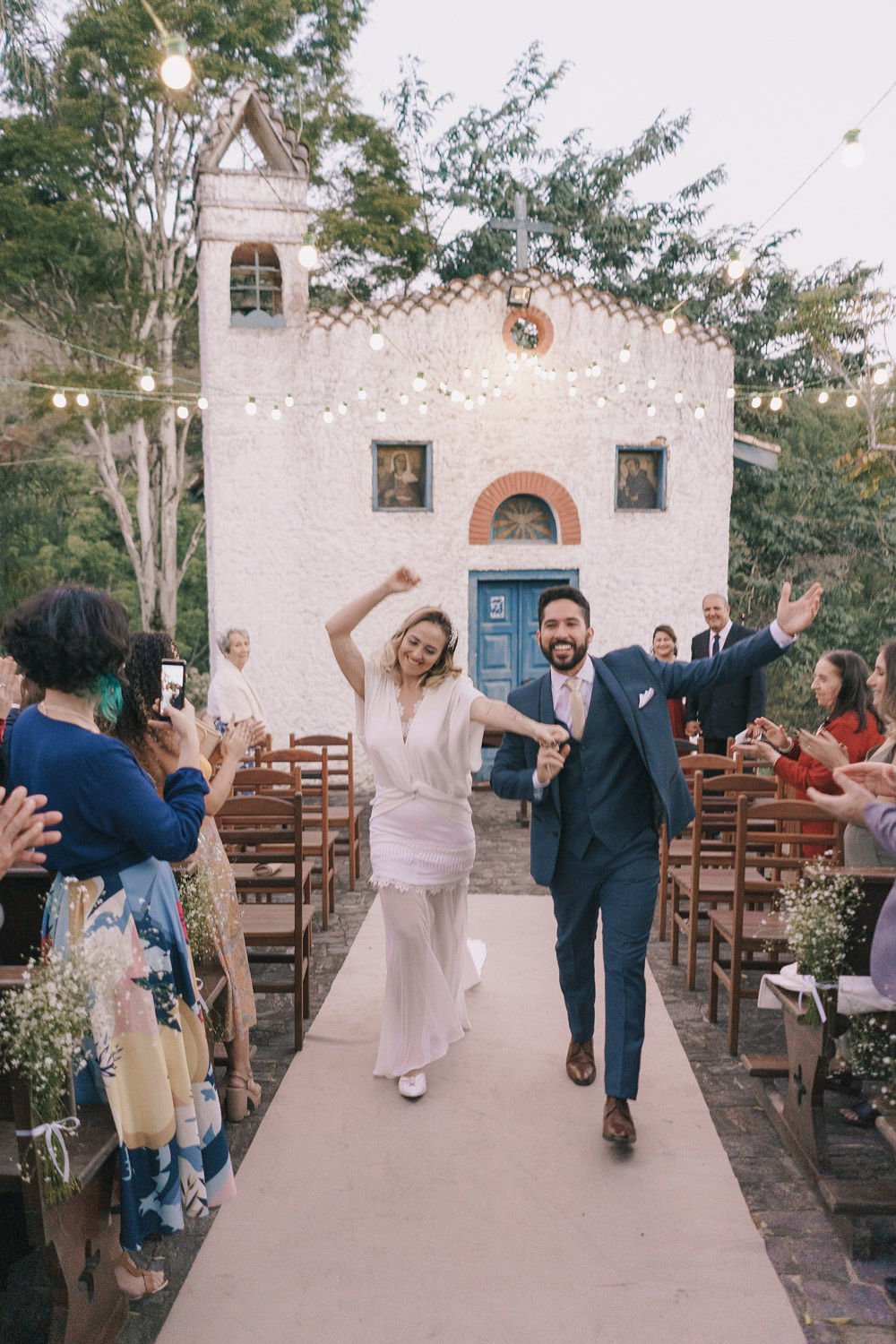 Casal de casamento caminhando pelo corredor na frente de uma igreja rústica com decoração de luzes e convidados aplaudindo.