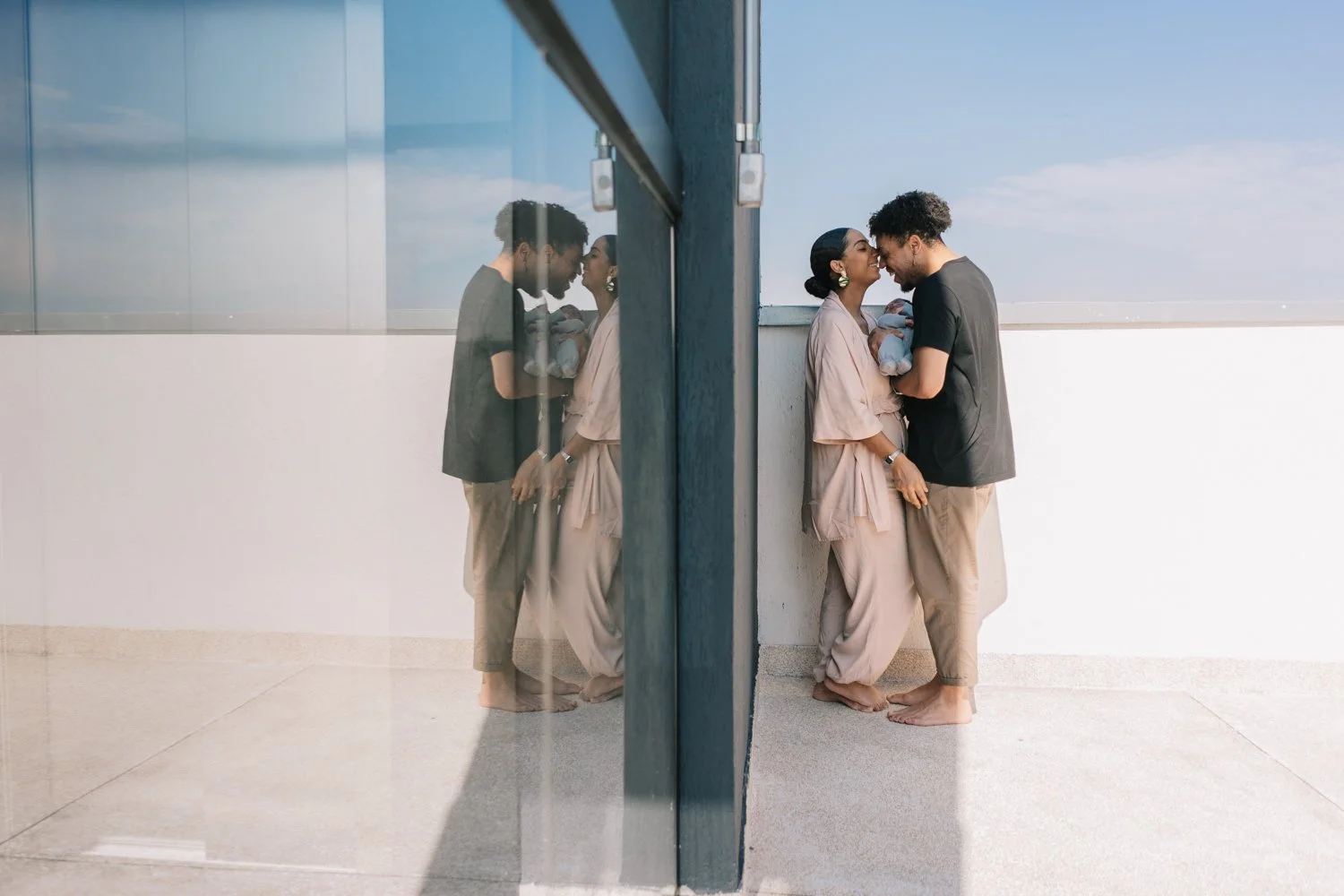 Casal se abraçando e sorrindo na varanda enquanto segura um bebê, com reflexão visível na porta de vidro ao lado.