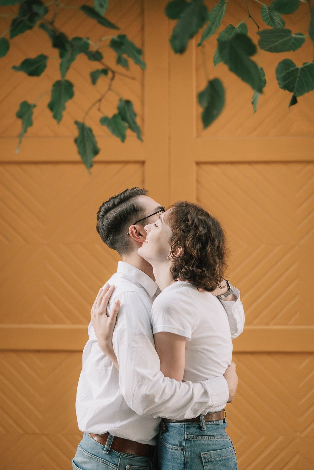 Casal jovem se abraçando e se beijando, com fundo de parede de madeira e folhas verdes penduradas.
