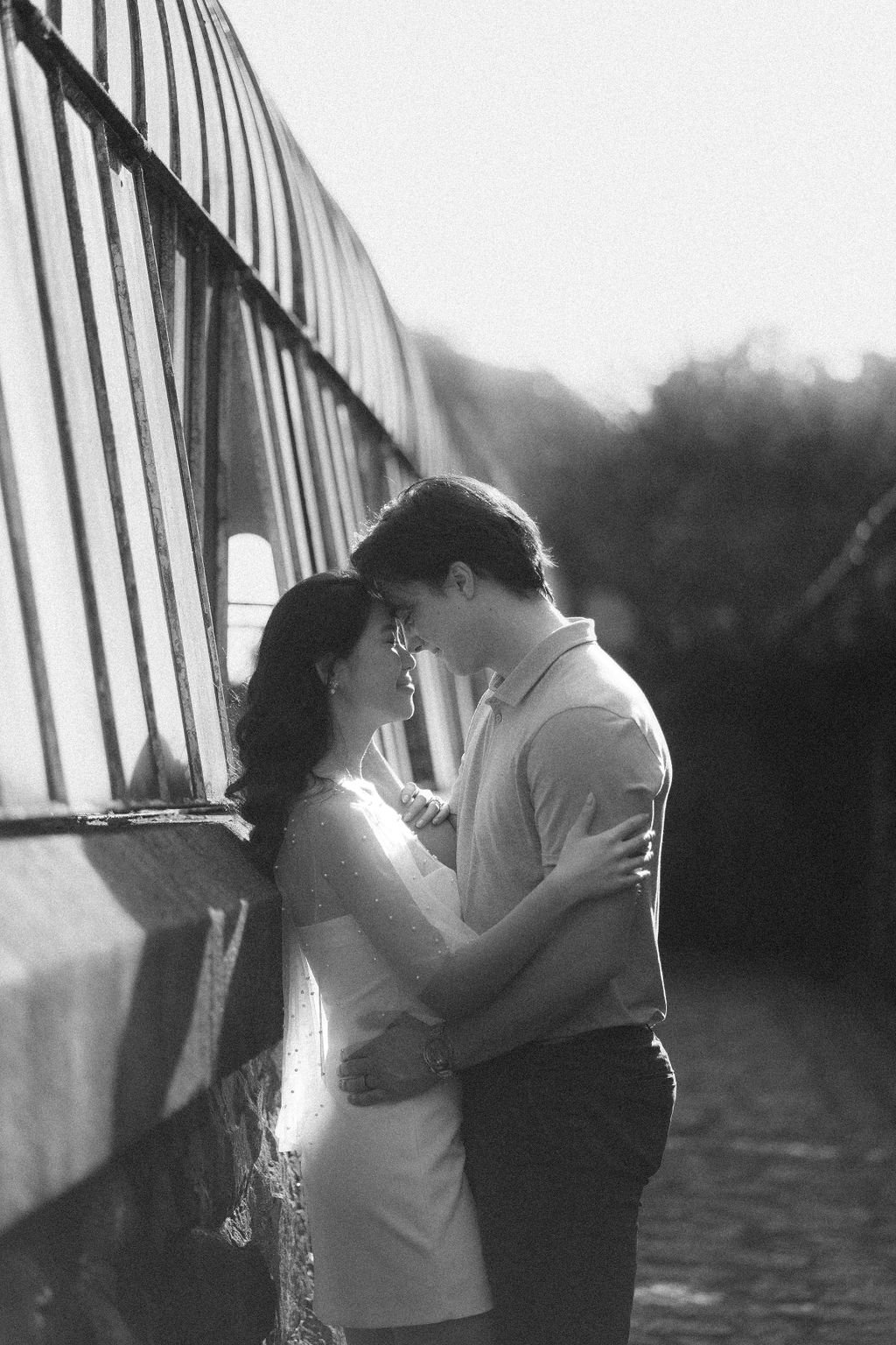 Um casal de jovens se envolvendo romanticamente, com a testa e o nariz juntos, em um momento de intimidade ao ar livre, ao pôr do sol, ao lado de uma cerca de ferro.