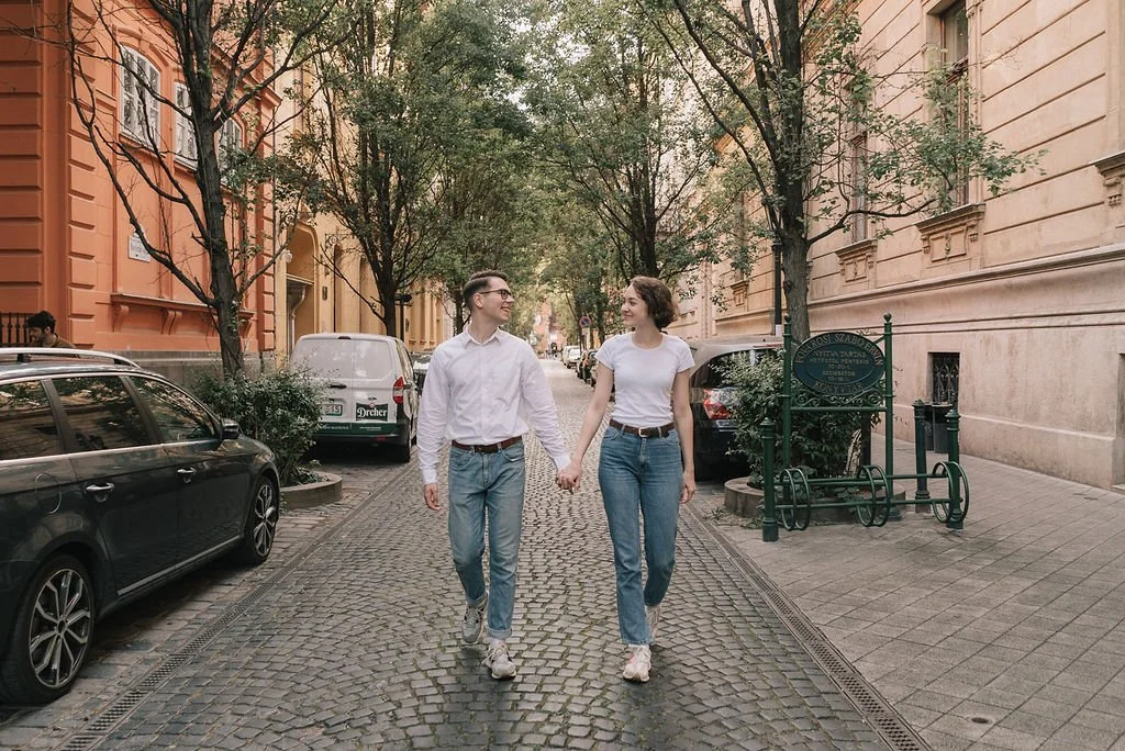 Homem e mulher caminhando de mãos dadas na rua, com carros estacionados ao lado e árvores ao fundo, em uma área urbana.