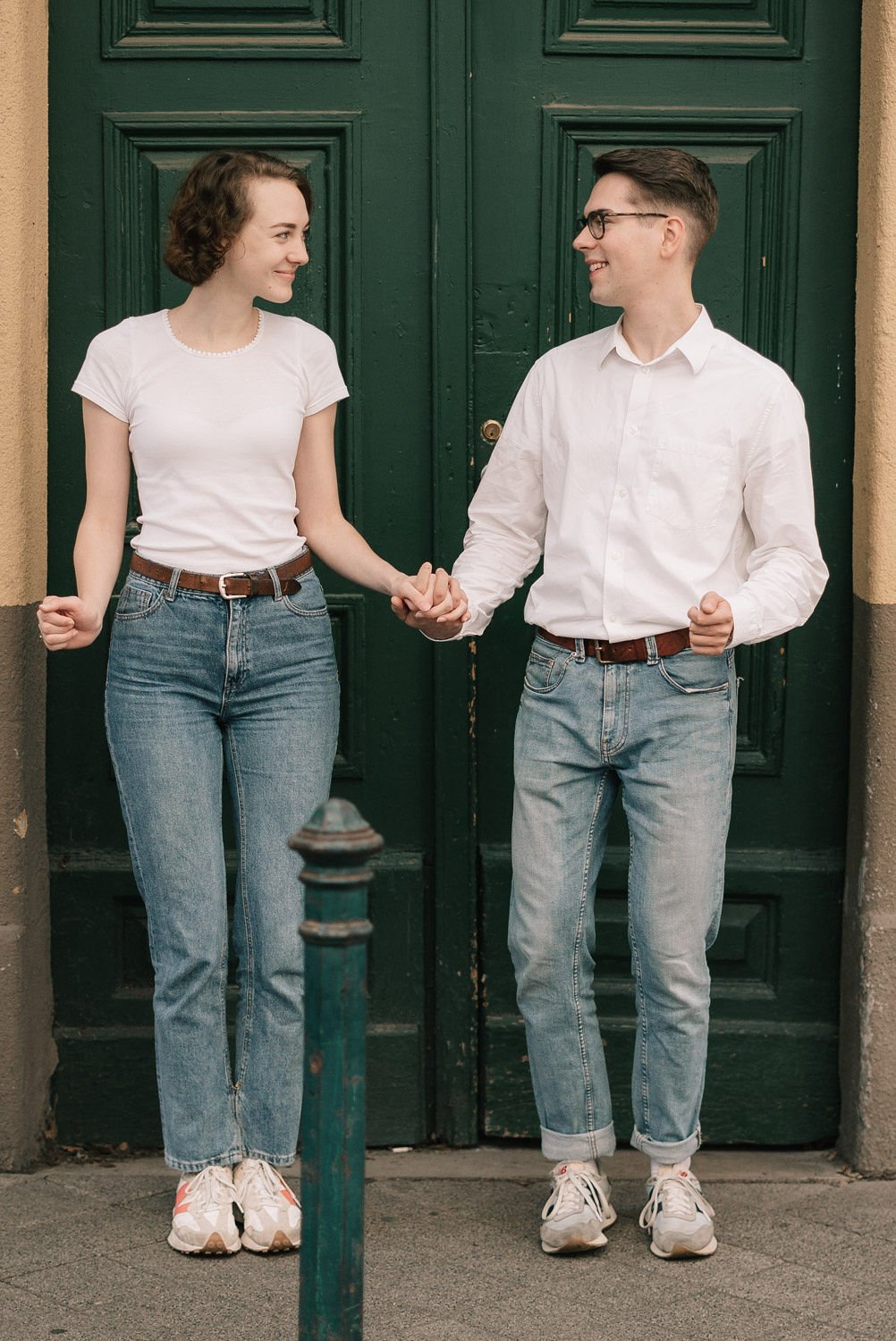 Um casal sorri e segura as mãos em frente a uma porta verde, usando roupas casuais. Ela tem cabelo cacheado curto, camiseta branca e jeans. Ele tem cabelo curto, óculos, camiseta branca e jeans.