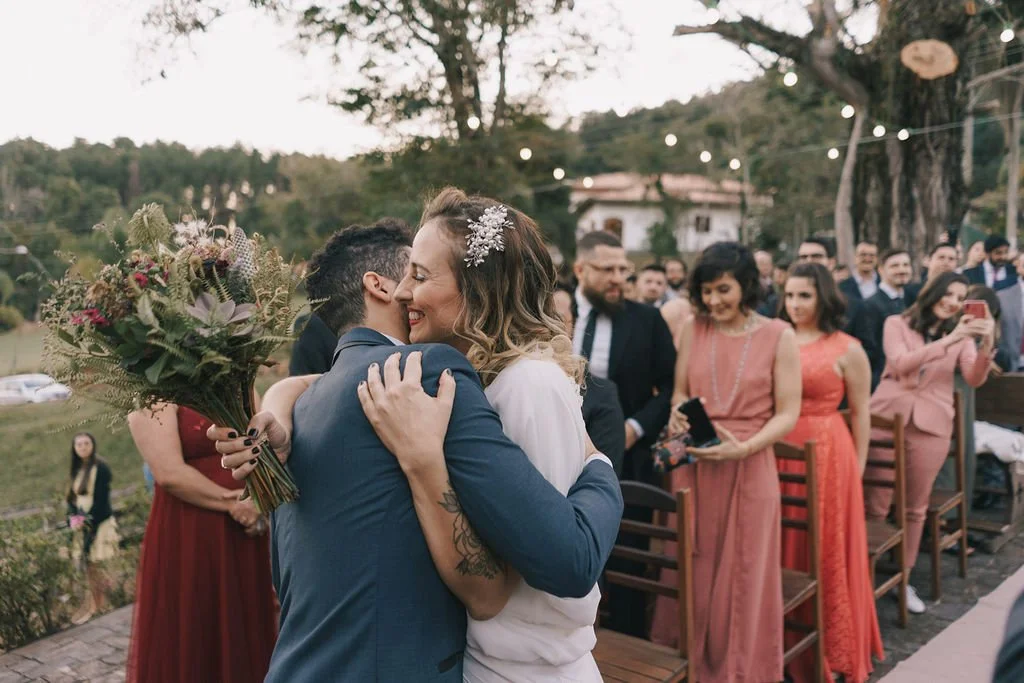 Casal se abraçando durante casamento ao ar livre, com convidados ao fundo e luzes decorativas.