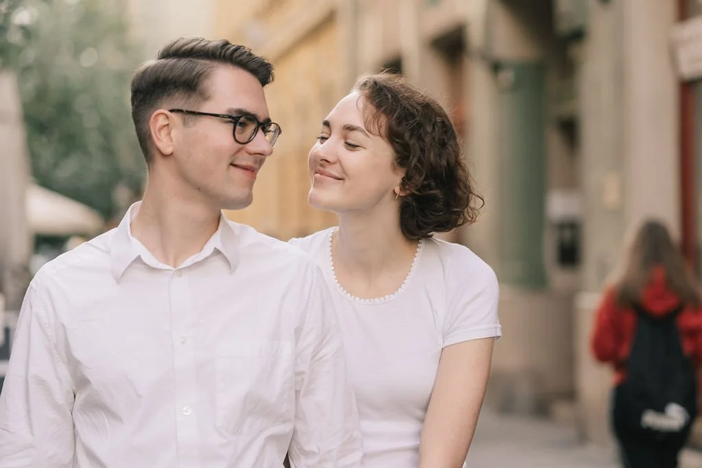 Casal sorridente caminhando na rua de cidade, ambos vestindo branco, com prédios ao fundo, mulher com cabelo curto cacheado e homem de óculos.