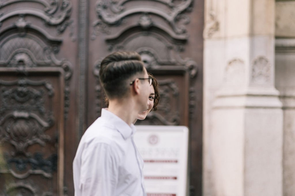 Homem com cabelo curto e óculos de sol, vestindo camisa branca, ao lado de uma mulher de cabelo cacheado, na frente de uma porta de madeira escura com detalhes intricados.
