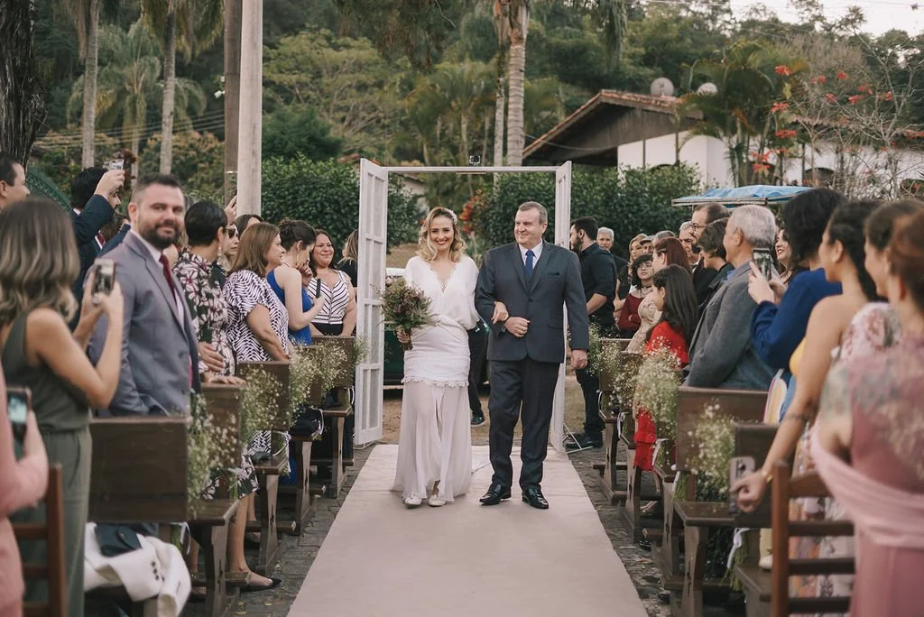 Casamento ao ar livre com a noiva de mãos dadas com o pai na saída, rodeados por convidados felizes