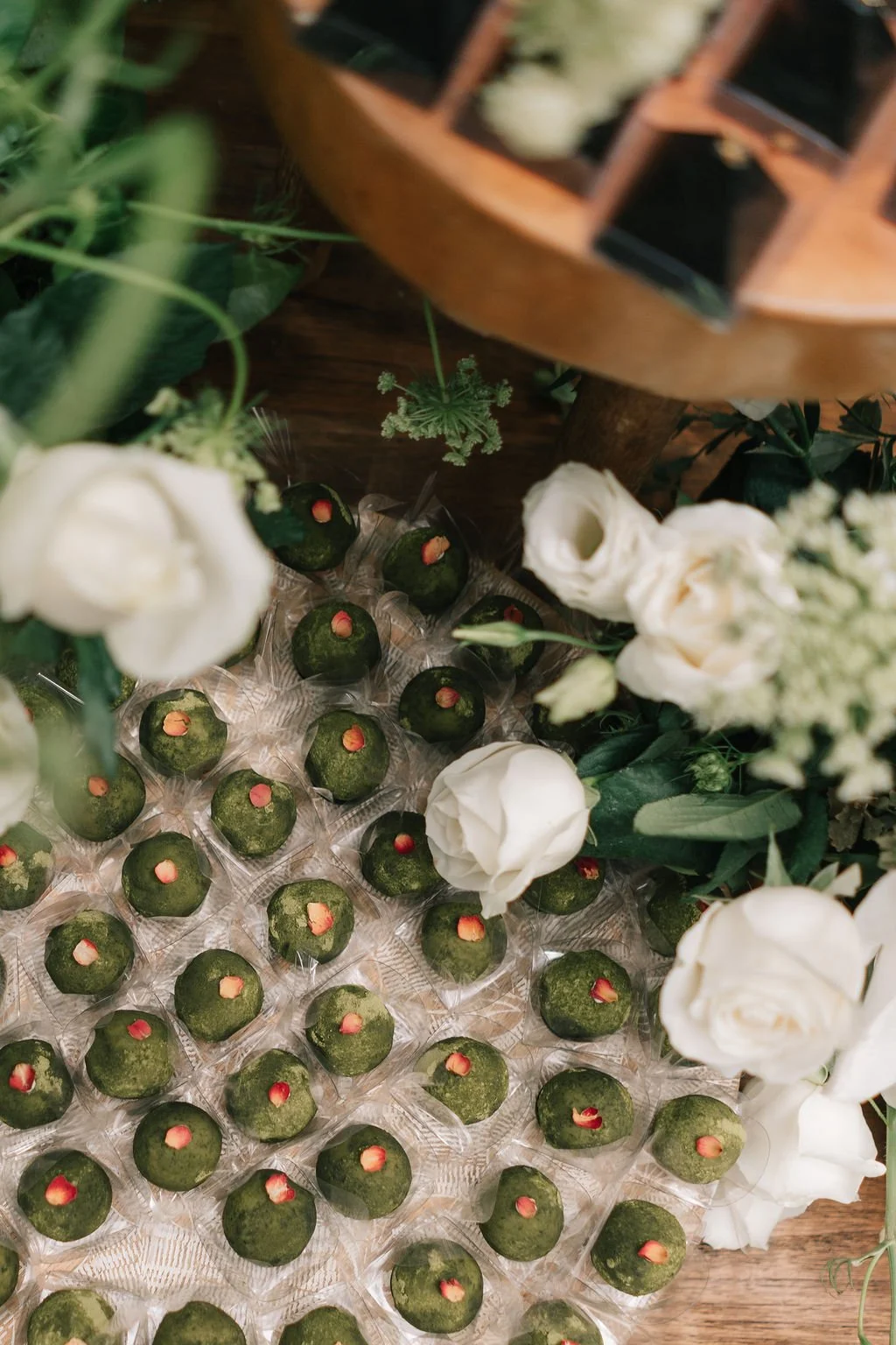 variedade de doces verdes, decorados com pétalas de rosa, cercados por flores brancas e folhagens verdes em uma mesa de madeira