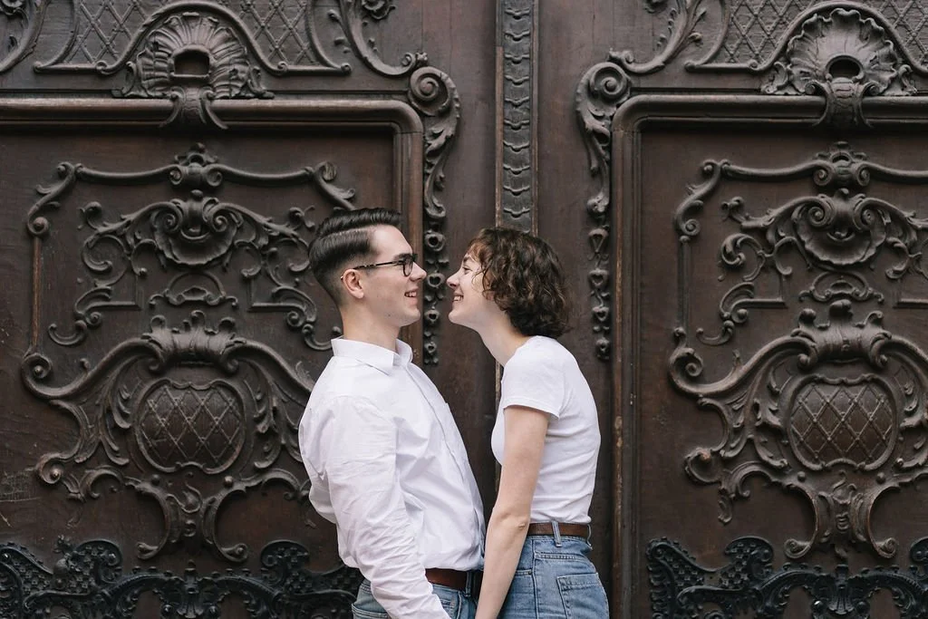 Um casal jovem sorrindo, de frente um para o outro, em frente a uma grande porta de madeira com detalhes ornamentais.