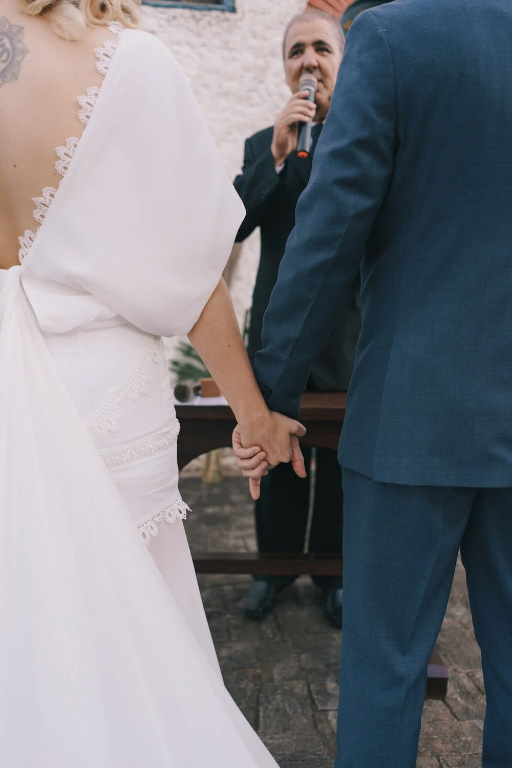 Noivos de mãos dadas durante cerimônia, com um homem com microfone ao fundo