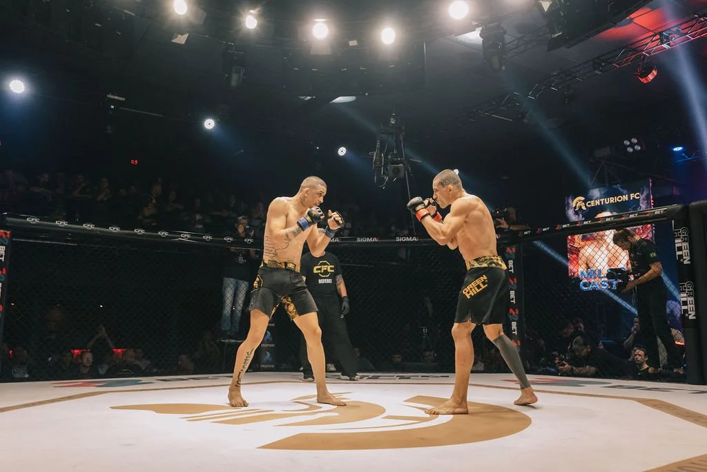 Dois lutadores de MMA em combate dentro de uma jaula de octógono na noite, com público ao fundo e câmeras. Ambos usam shorts e estarão se enfrentando.