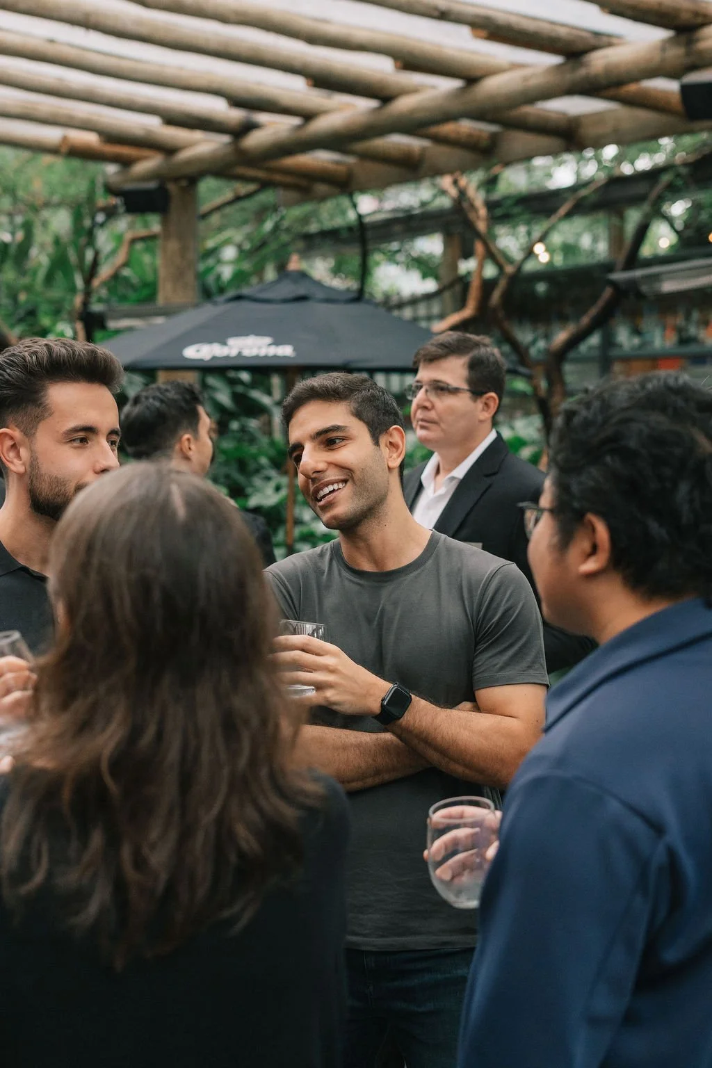 Grupo de pessoas conversando em um ambiente ao ar livre com árvores e uma estrutura de madeira no fundo, alguns segurando copos de bebida.