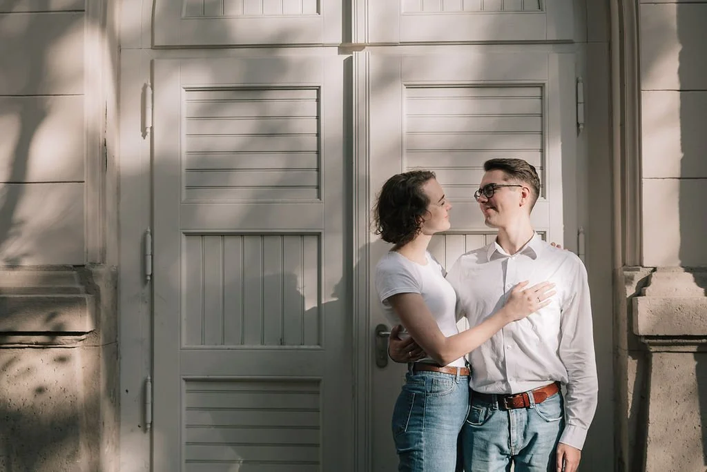 Casal sorridente olhando-se em frente na porta de entrada de uma casa, com sombra de árvore ao fundo.