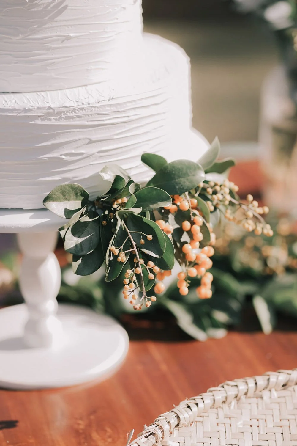 Bolo branco decorado com folhas verdes e frutas laranja, em uma festa ao ar livre.