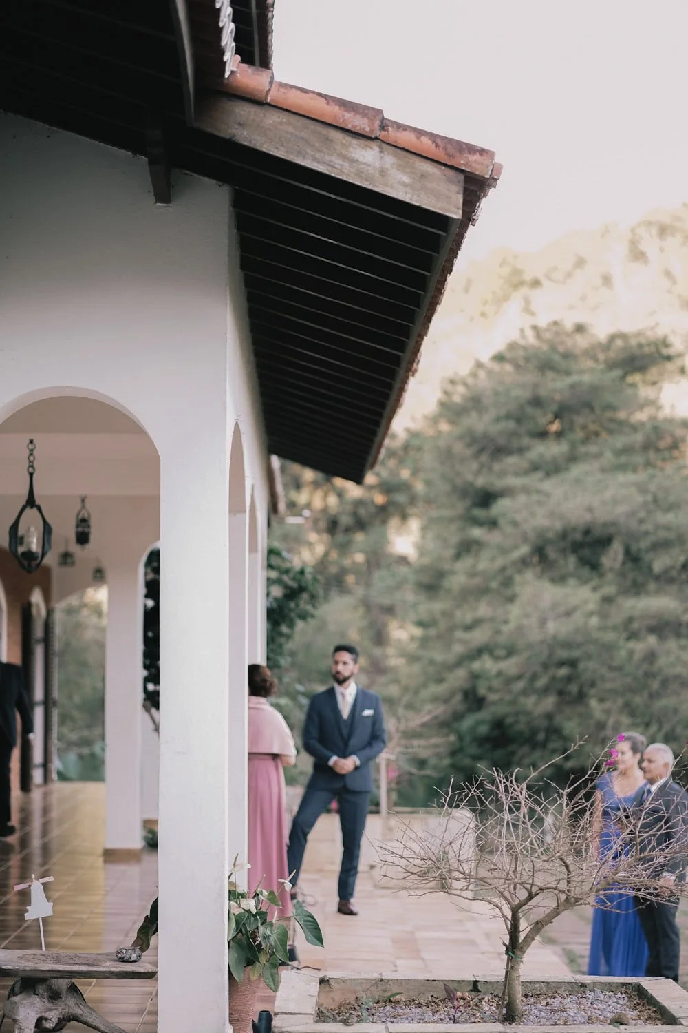 Pessoas conversando na varanda de uma casa com jardim, árvores ao fundo e iluminação de luz quente.
