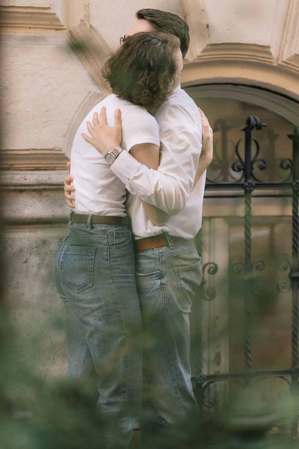 Dois adultos abraçando-se, vistos de costas, em frente a uma parede de pedra com uma janela arqueada e grades de ferro.