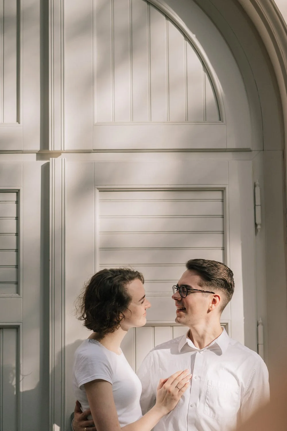 Casal sorrindo se olhando em frente a uma porta branca com detalhes decorativos.