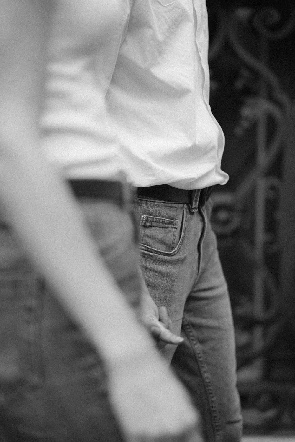 Pessoa com calça jeans e camisa branca, segurando a mão na cintura, ao lado de outra pessoa, em ambiente com fundo decorativo.