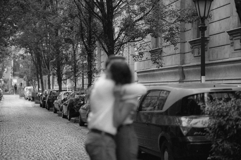 Rua com calçada de paralelepípedos, vários carros estacionados ao longo da via, árvores alinhadas ao lado da calçada e uma pessoa de cabelo curto caminhando com uma mochila. Edifícios de arquitetura clássica e uma lâmpada de iluminação pública também