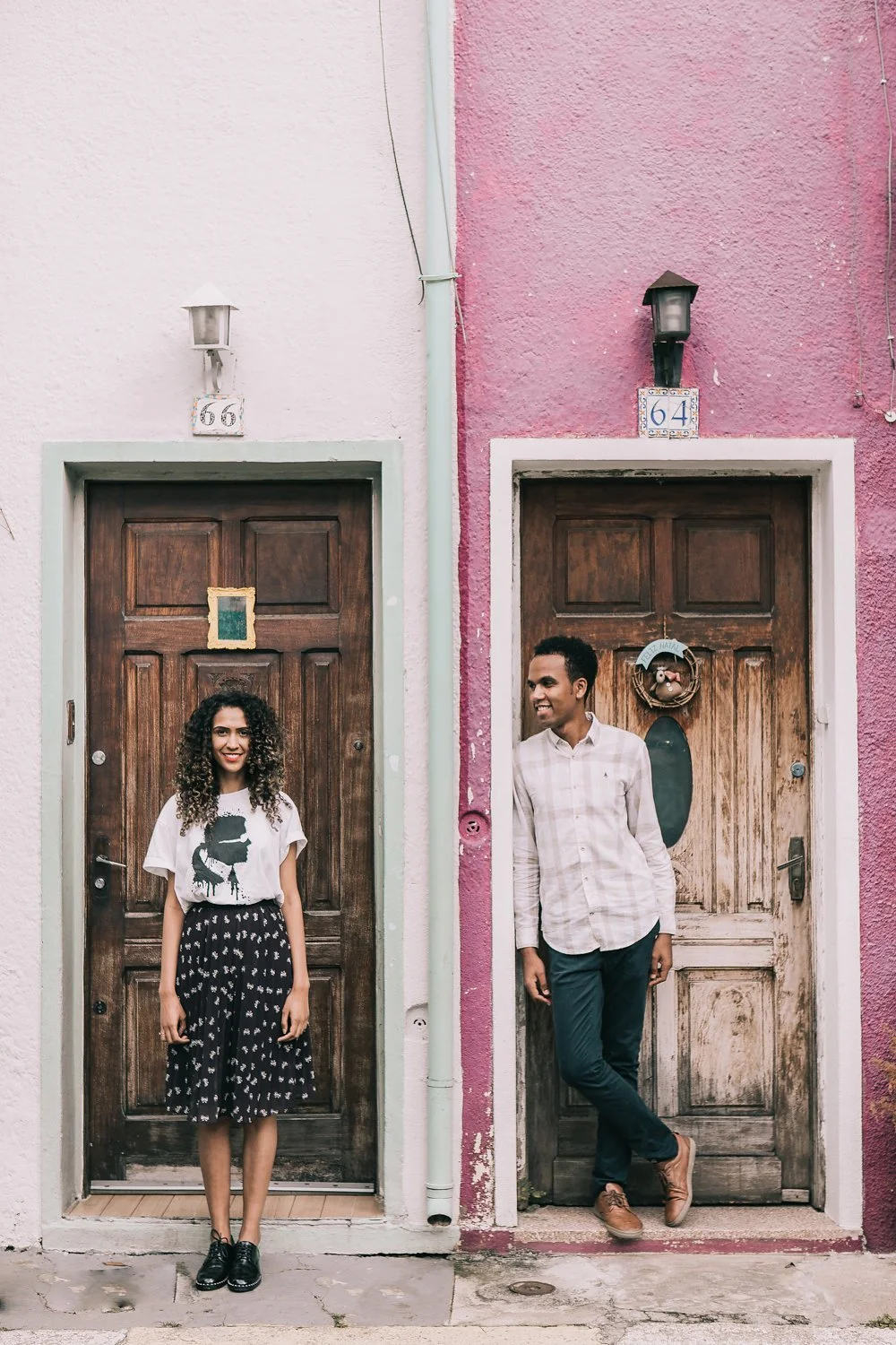 Um homem e uma mulher jovens, sorrindo, posando em frente a portas de madeira em casas coloridas, uma rosa e outra branca, com números 66 e 64 acima das portas.