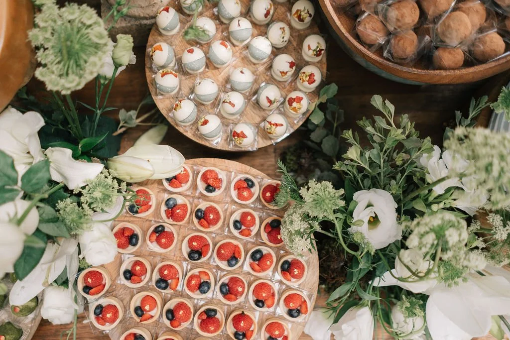 Prato de sobremesas com morangos, amoras e outras frutas em copinhos de vidro, acompanhado de flores brancas e verdes e doces em uma bandeja de madeira.