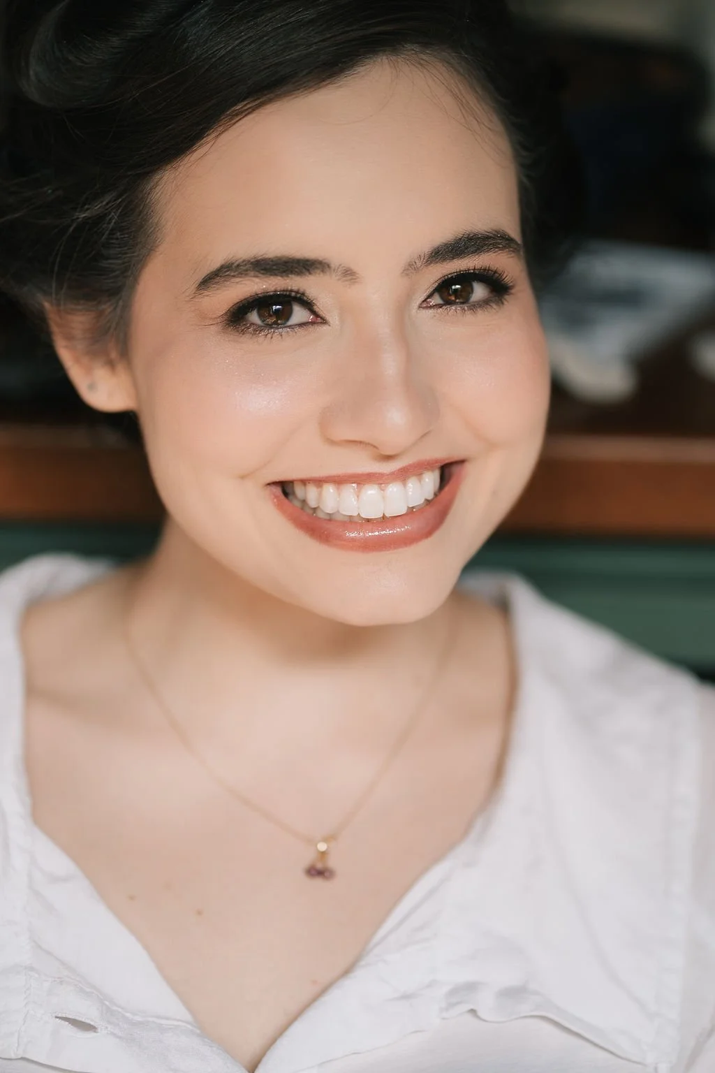Retrato de uma mulher sorrindo, com pele clara, cabelo escuro e olhos castanhos, usando uma blusa branca e um colar delicado.