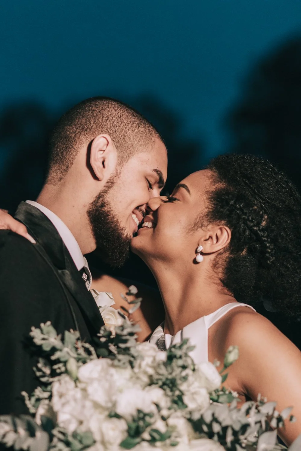 Casal de noivos se beijando em comemoração de casamento, com buquê de flores na frente.