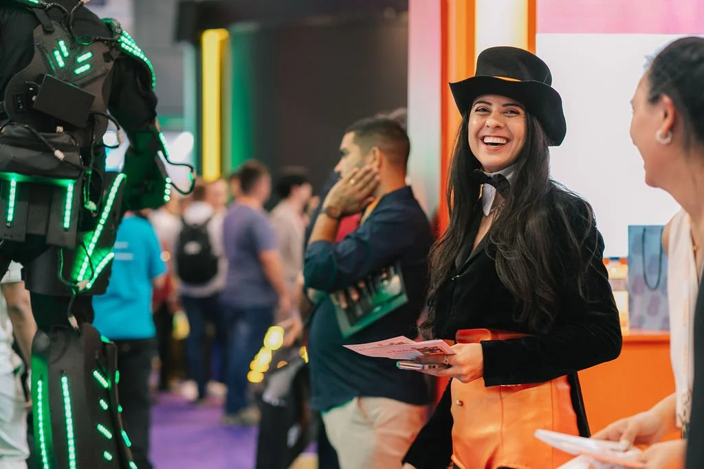 Mulher sorridente usando chapéu preto e roupa de show ou evento, conversando com outra pessoa, com uma fila de pessoas ao fundo em ambiente de evento ou conferência.