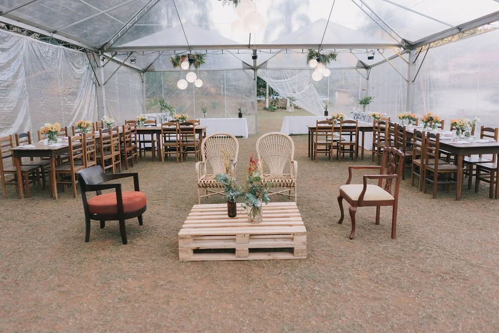 Decoração de evento ao ar livre com mesas e cadeiras sob uma tenda transparente, incluindo cadeiras de madeira, poltronas de vime, uma mesa de pallets com arranjos florais, iluminação suspensa e uma área decorada com tecidos brancos ao fundo.